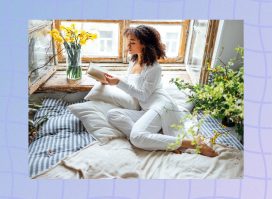 focused woman wearing white PJs and white robe reading a book in bed in front of window surrounded by flowers