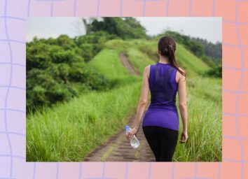 back of brunette woman wearing purple tank top and black leggings walking on dirt hiking path surrounded by tall grass