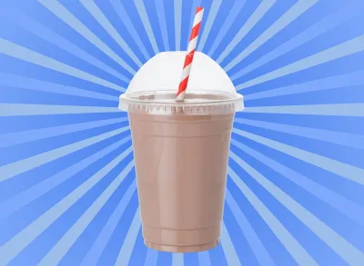 A chocolate milkshake in a clear plastic cup with a striped straw set against a vibrant blue background