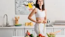 Selective focus of fit woman smiling at camera while measuring waist with tape near salad and fresh vegetables on table.