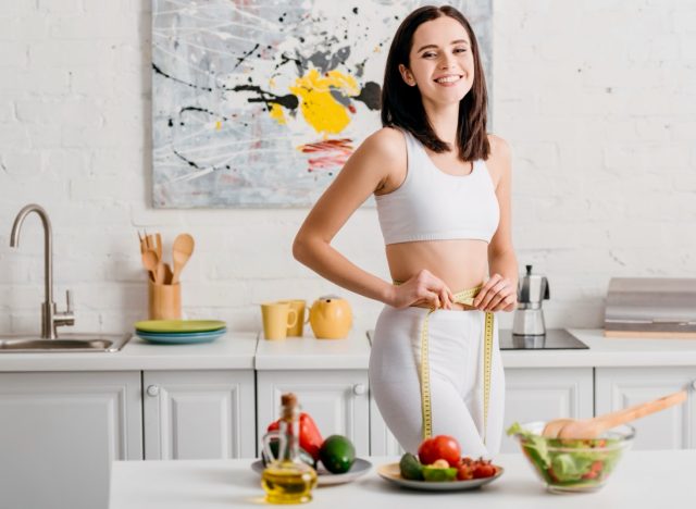 Selective focus of fit woman smiling at camera while measuring waist with tape near salad and fresh vegetables on table.