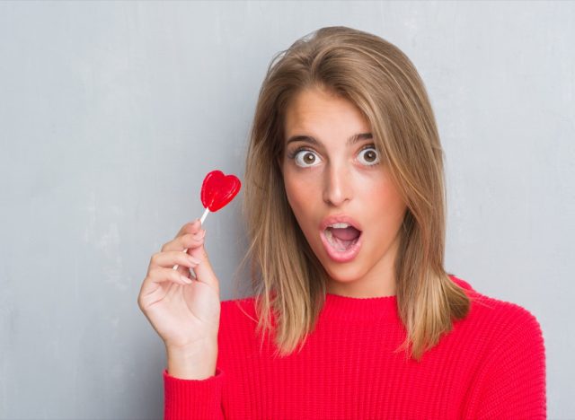 Beautiful young woman over grunge grey wall eating red heart lollipop candy scared in shock with a surprise face, afraid and excited with fear expression