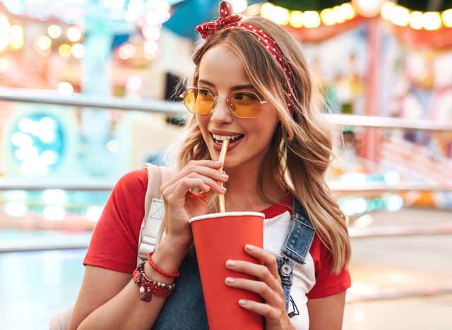 Image of attractive charming woman wearing girlish clothes drinking soda beverage from paper cup while walking in amusement park