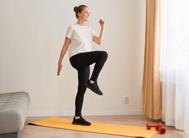 Full length portrait of slim athletic woman wearing white t shirt and black leggins doing sport exercises at home on yoga mat, walking in place, marching, sport, fitness at home.