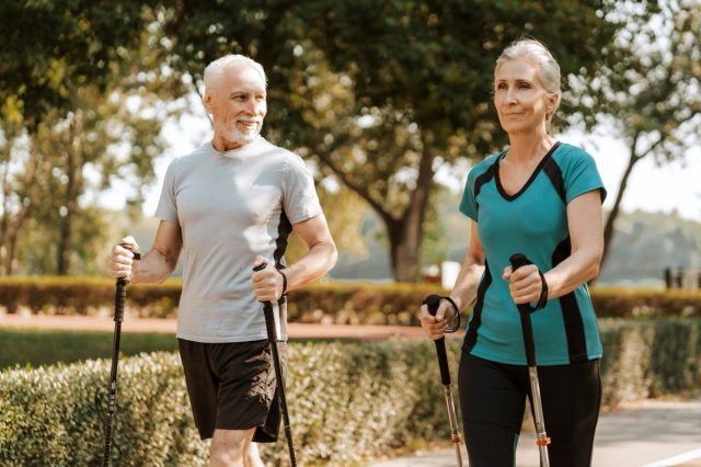 Senior man and woman are nordic walking on a trail outside on a sunny day. They are both wearing athletic wear and are smiling. Active lifestyle concept