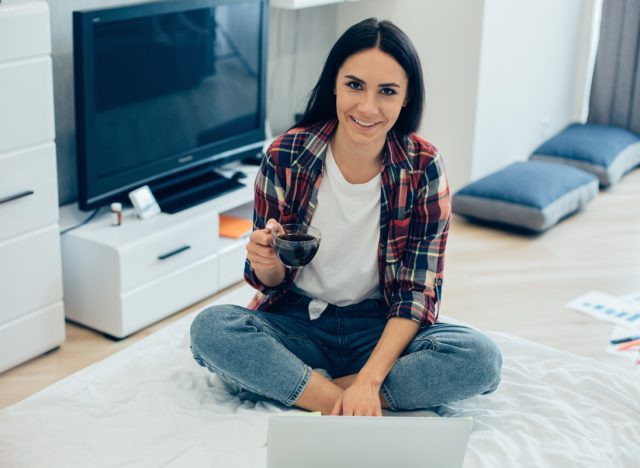 Positive smiling young woman sitting cross-legged on the bed in front of a laptop and holding a cup of coffee