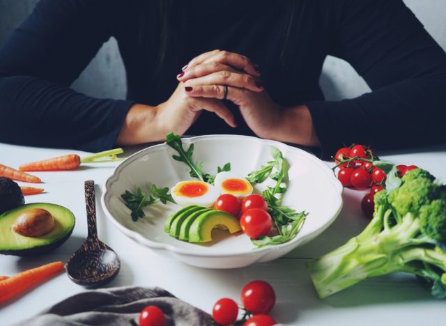 Weight loss plans for women theme white ceramic bowl contained healthy salad (slided avocados, boiled eggs, rocket leaves, tomatoes) on wooden table with blurred woman in black dress / selective focus