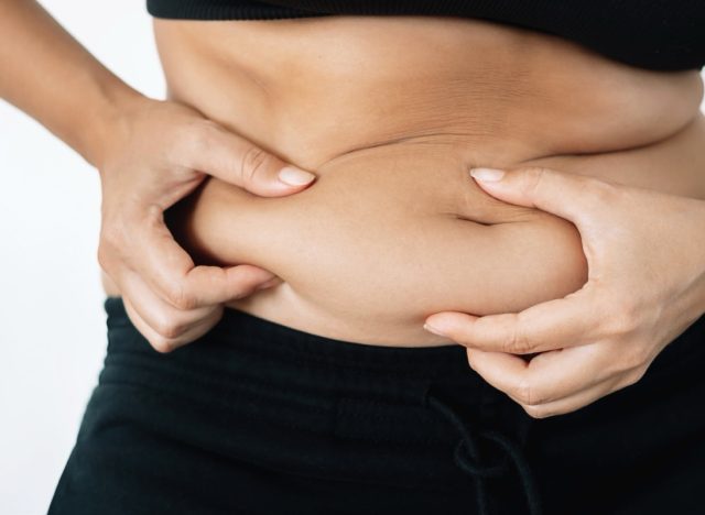 A cropped shot of a young woman in a black underwear holding herself by the fat on her stomach isolated on a white background. Overeating, overweight, excess weight, hormonal belly, rolls