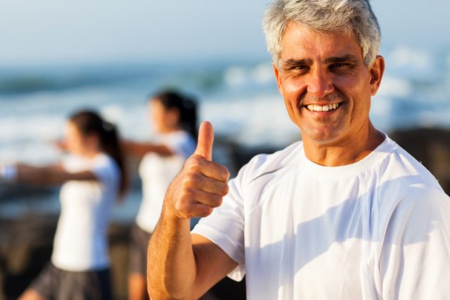 Active mature man giving thumb up on beach with family exercising on background.