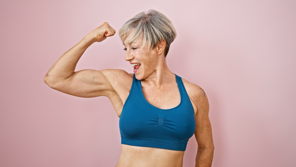 A fit, mature woman with short grey hair flexes her muscles against a pink isolated background, displaying strength and vitality.