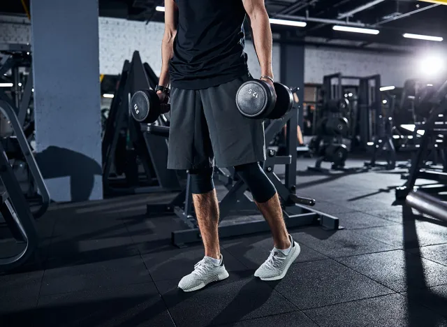 Cropped photo of a sportsman in shorts and white boots walking away with hand weights, farmer's carry, farmer carry