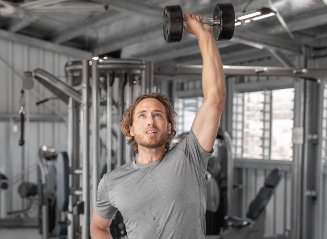 Gym workout fitness man training with dumbbells lifting overhead raise shoulders exercise shoulder press indoors at health club. Square crop.