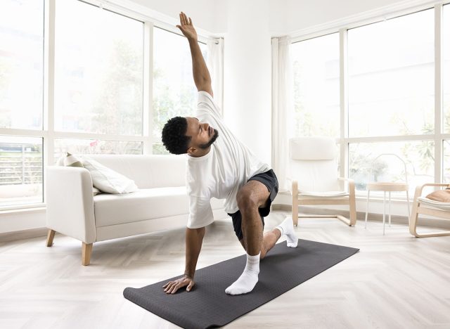 Serious young Black man doing morning exercises at home, keeping twisting yoga asana on mat, caring for wellness, fit, wellbeing, healthy active lifestyle, training body in apartment