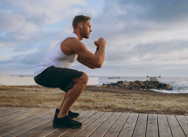 Full body young strong sporty athletic toned fit sportsman man wears sports clothes earphones do squats warm up train at sunrise sun dawn over sea beach outdoor on pier seaside in summer day morning