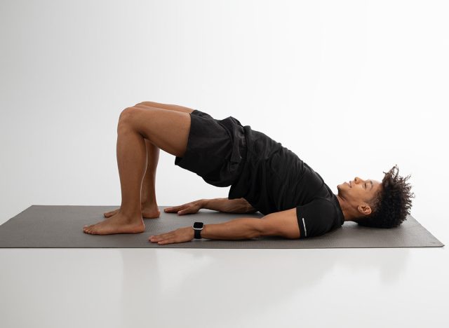 A man is engaging in the bridge exercise on a yoga mat in a well-lit indoor studio. He emphasizes strength and stability as he raises his hips, focusing on core and glute engagement