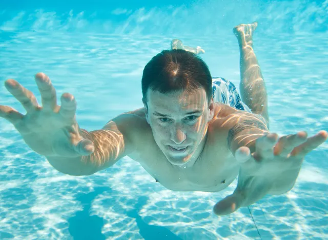 Man swimming with open eyes underwater in pool, holding breath.