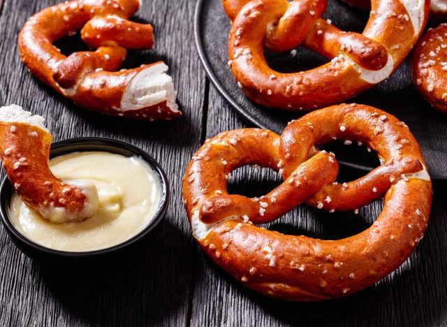 close-up of soft pretzels baked in the form of knot and sprinkled with salt on black plate on dark wooden table with cheese sauce, horizontal view from above