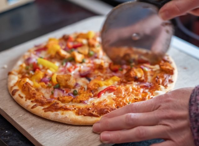 Close up and selective focus of a home cooked thin crust sweet chilli chicken pizza on a wooden cutting board being sliced with a pizza roller by an unidentifiable woman