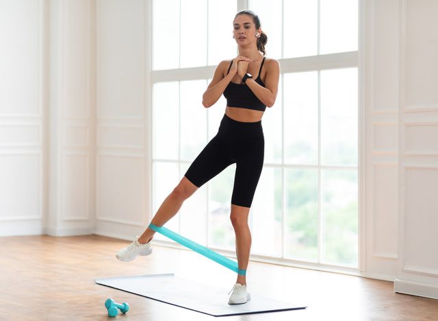 Full And Lower Body Workout. Focused Sporty Woman Exercising With Elastic Rubber Band Lifting And Kicking Leg Aside Standing On Yoga Mat, Listening To Music In Wireless Earpods. Bodyweight Training. Standing leg lifts.
