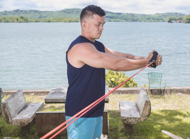 A fit middle aged man does banded low to high wood chops. A vacationer training outside at a lakeside park or resort.