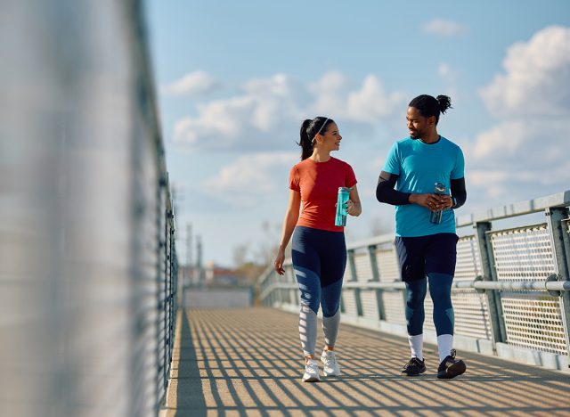 Happy athlete and her friend drinking water and communicating while walking over the bridge outdoors. 