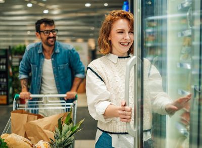 Young redhead woman opening refrigerated section door, smiling, choosing frozen food, while her boyfriend pushing shopping cart in supermarket