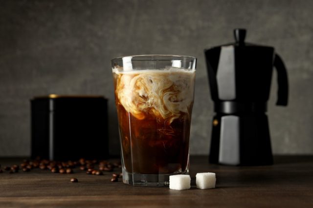 Glass with iced coffee drink, coffee maker, coffee bean and sugar on gray background
