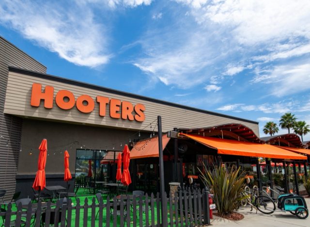 Long Beach, CA, USA: 2024 August 01: Hooters restaurant store front entrance exterior, logo, sign, sunny, entrance blue sky and clouds, green landscape.