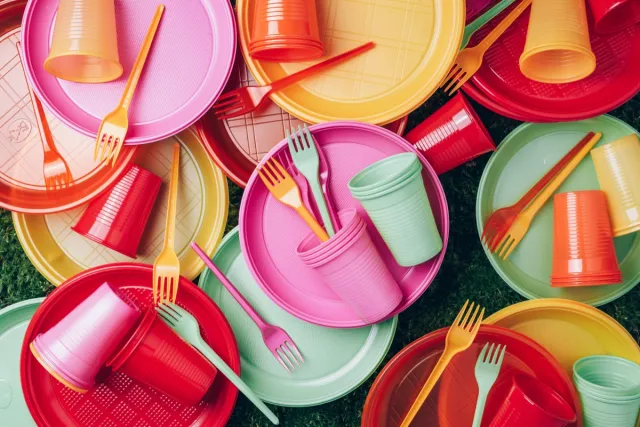 Disposable plastic tableware - plates, forks, spoons. Colored plastic disposable tableware on green grass moss background. Top view. Copy space. Birthday picnic utensil. Recycling plastic and ecology.