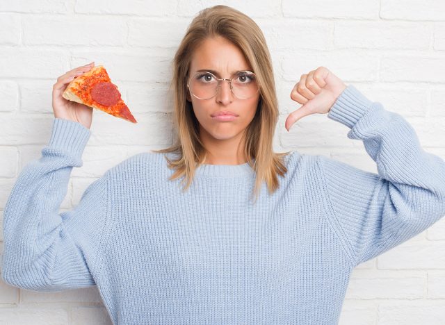 Beautiful young woman over white brick wall eating pizza slice with angry face, negative sign showing dislike with thumbs down, rejection concept