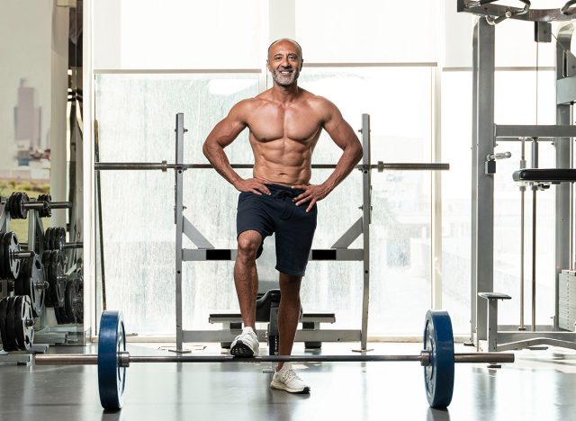 A strong muscular shirtless mature older bodybuilding athlete with balding gray hair standing with one foot on a heavy barbell, smiling at the camera in a gym, standing on one foot 