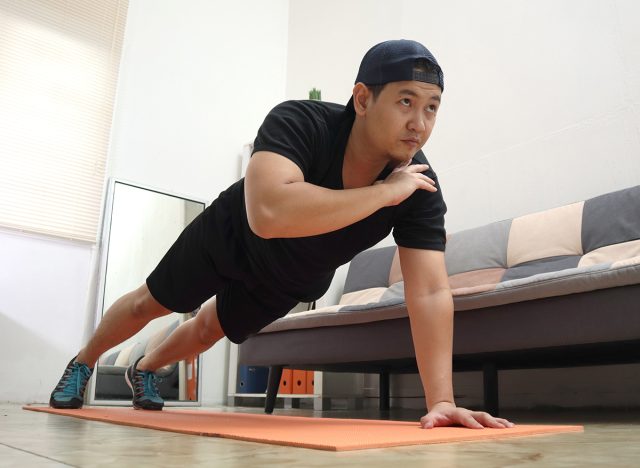 Asian male doing exercise at home to stay healthy on new normal lifestyle, indoor home workout concept, shoulder tap push ups plank position