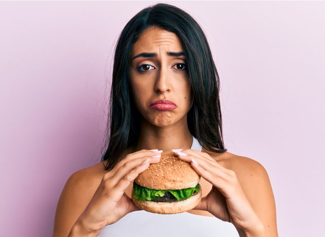 Beautiful hispanic woman eating a tasty classic burger depressed and worry for distress, crying angry and afraid. sad expression.