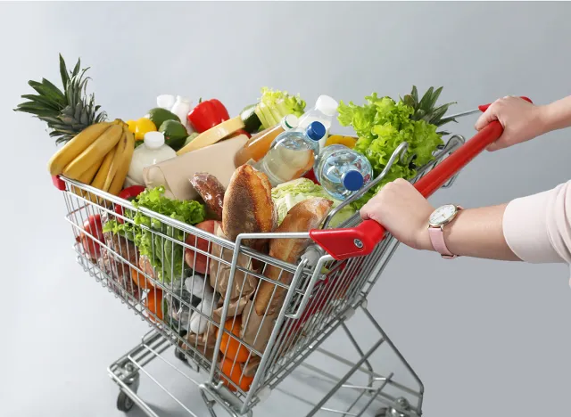 Woman with shopping cart full of groceries on grey background, closeup