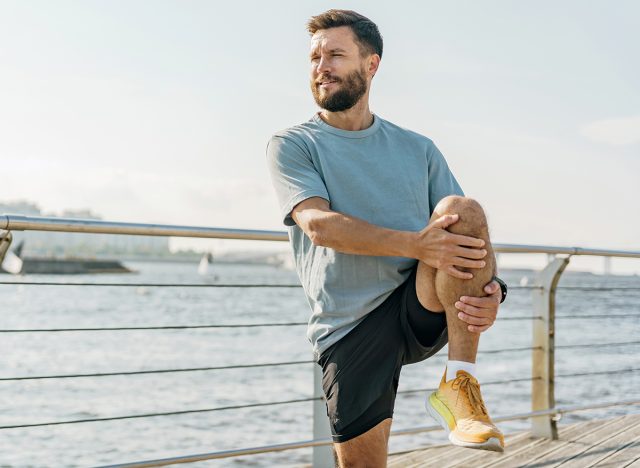 A focused man stretches his leg on a sunlit boardwalk by the river, gazing into the distance, contemplating his workout.