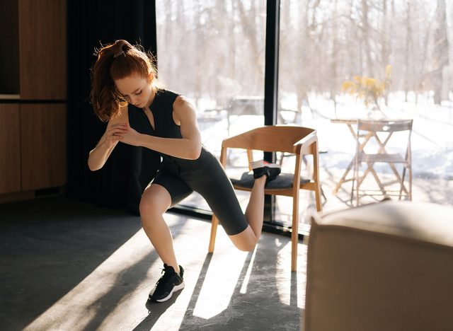 Sportive slim female doing single leg lunge using chair in living room. Front view of motivated fitness woman performing single leg lunge pose on chair. Home fitness and wellness concept