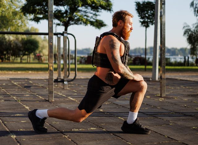 Bearded man in weighted vest training in park in morning, lunges 