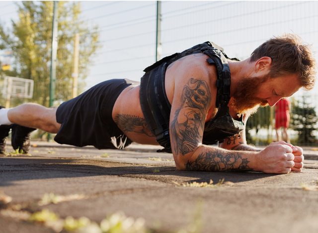 Tattooed sportsman doing plank in park in morning, weighted vest