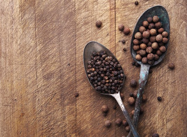 Image of two vintage spoons full of black peppercorn and allspice on rustic wooden cutting board