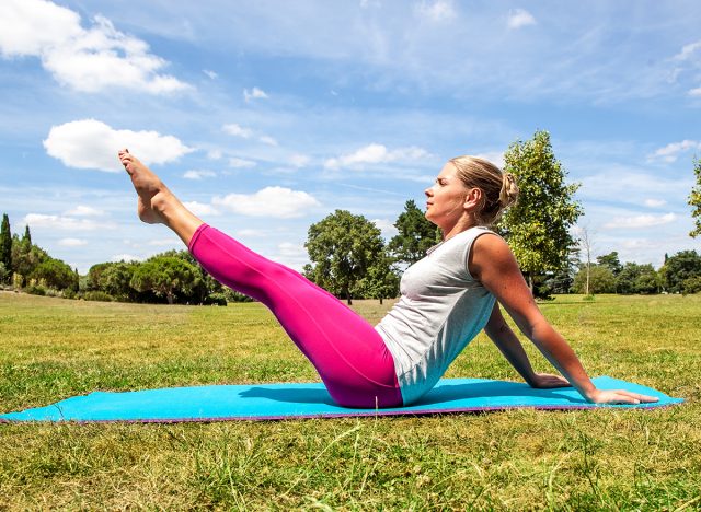 fitness and gym outside - active young blond woman working out to keep fit, exercising her toned legs and abs on an exercise mat over sunny blue sky, pilates, boat pose. 