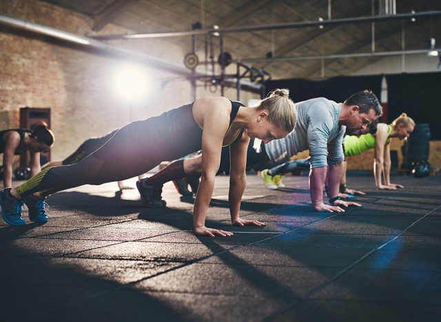 Group of adults doing push up exercises at indoor physical fitness cross-training exercise facility with bright light flare over them, extended straight arm plank 