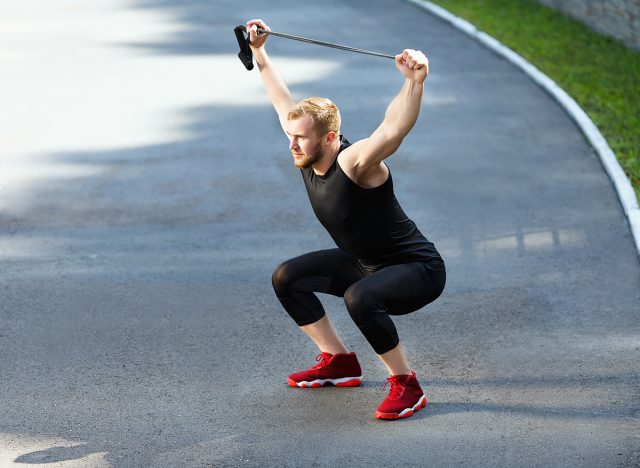 Profile of man training with expander. Muscular sportsman squatting with expander, hands up. Sport, outdoors, stadium, full body, horizontal, fast feet and reach 