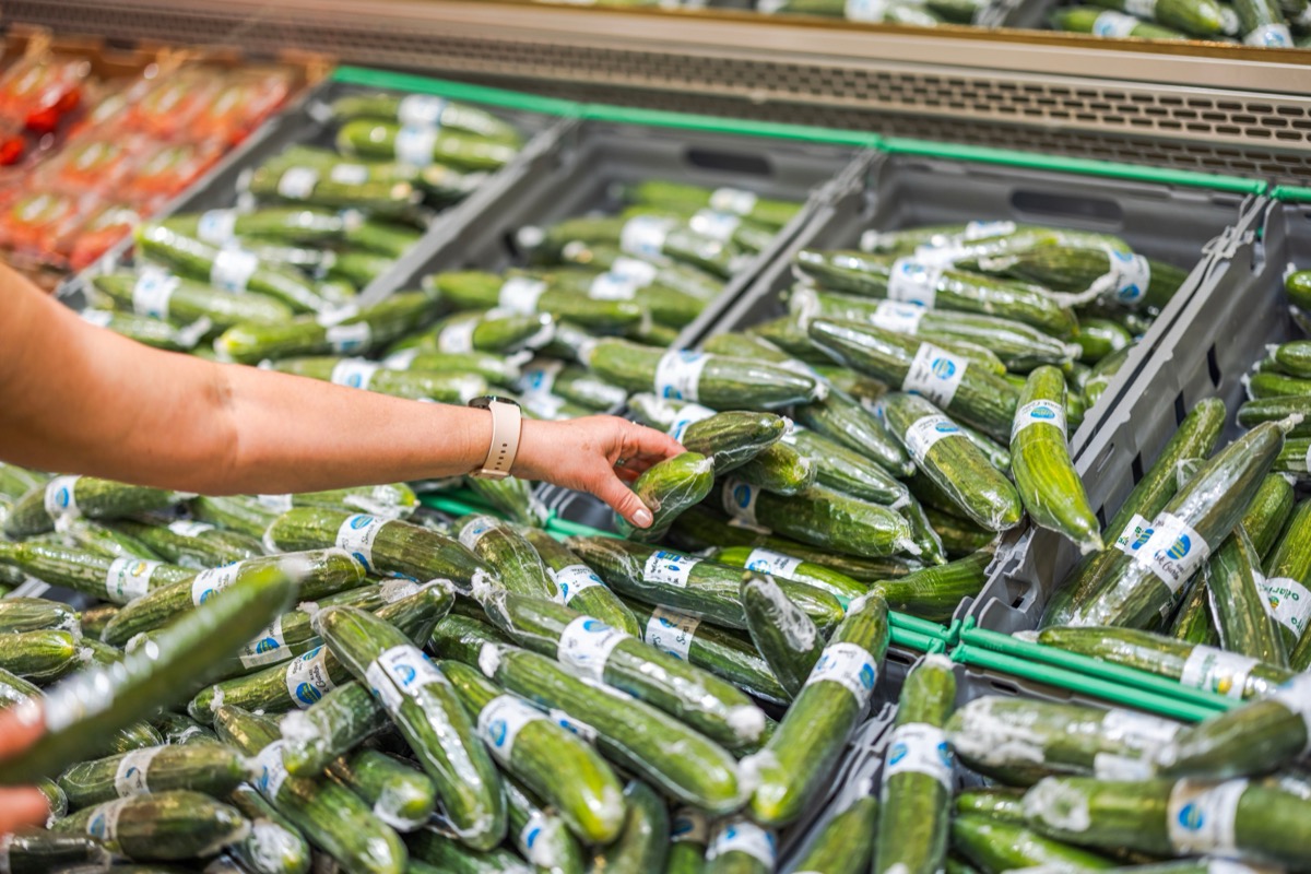 Close-up view of a hand picking up a cucumber wrapped in vacuum packaging from the shelf in the store.
