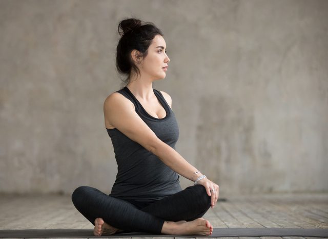 Young sporty woman practicing yoga, doing Revolved Easy exercise, Parivrtta Sukhasana pose, working out, wearing sportswear, black pants and top, indoor full length, gray wall, yoga studio. Seated Cross-Legged Stretch