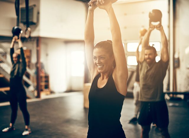 Smiling young woman in sportswear swinging a dumbbell over her head during a gym exercise class