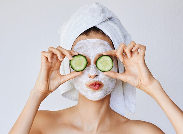 Beautiful young woman with facial mask on her face holding slices of fresh cucumber. Skin care and treatment, spa, natural beauty and cosmetology concept, over white background