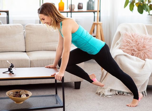 Sporty woman doing functional fitness exercise and watching online workout session on smartphone, training with coffee table in living room