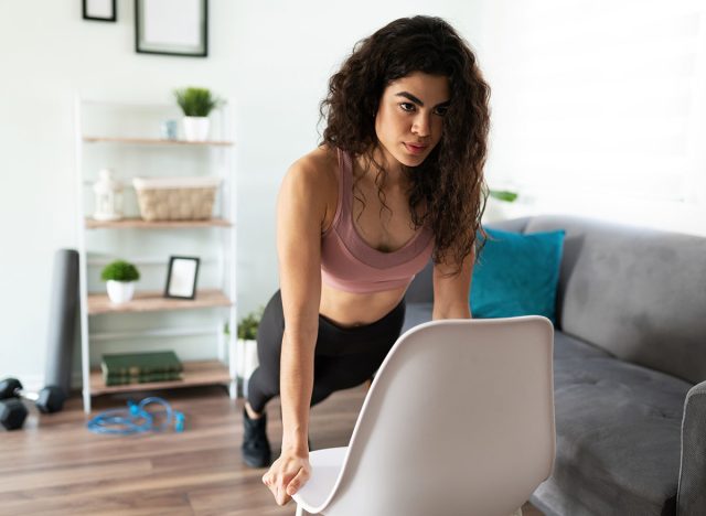 Front view of an attractive young woman doing push ups with a chair at her home living room