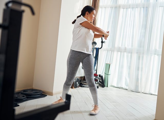 Full-length portrait of a serious well-built dark-haired Caucasian woman performing a standing trunk rotation exercise, twist 