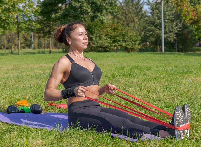 Muscular middle aged caucasian woman does seated row exercise for latissimus dorsi muscles using resistance band in city park on green grass. Black sportswear. Healthy lifestyle. Outdoor sport theme.